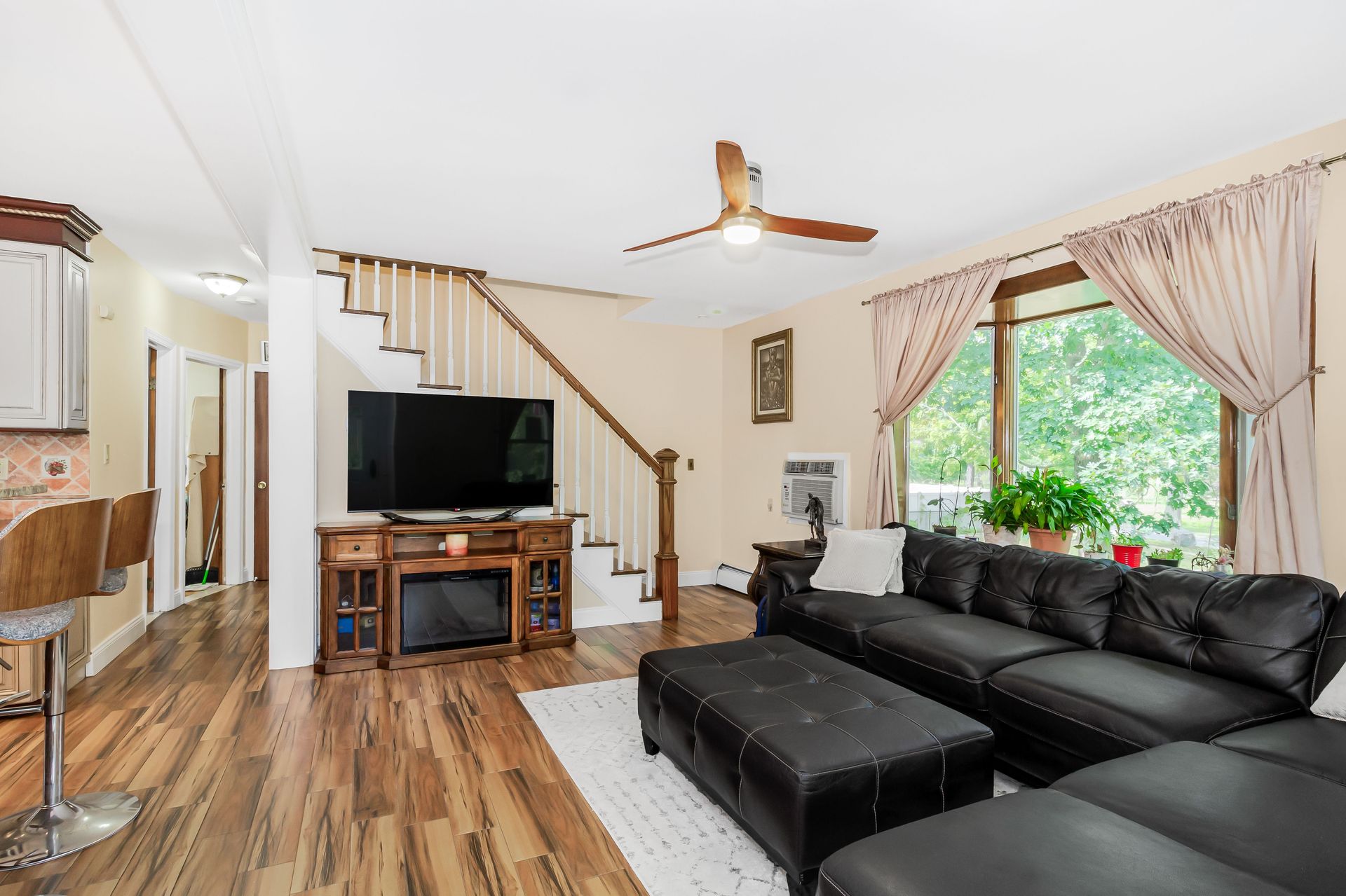 Bright living room with black leather sofa, TV, staircase, and window garden view.
